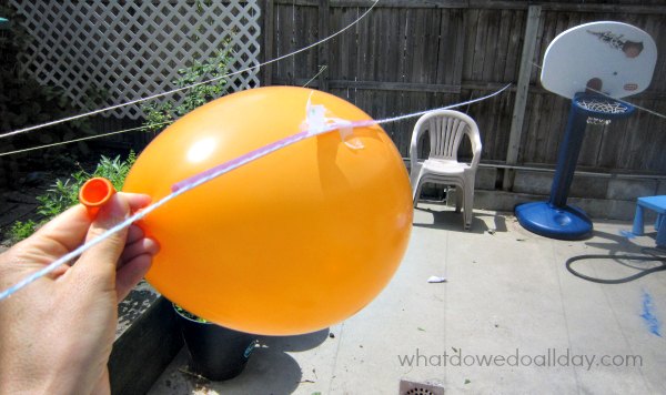 Hand holding orange balloon attached to string in backyard for a balloon rocket experiment.