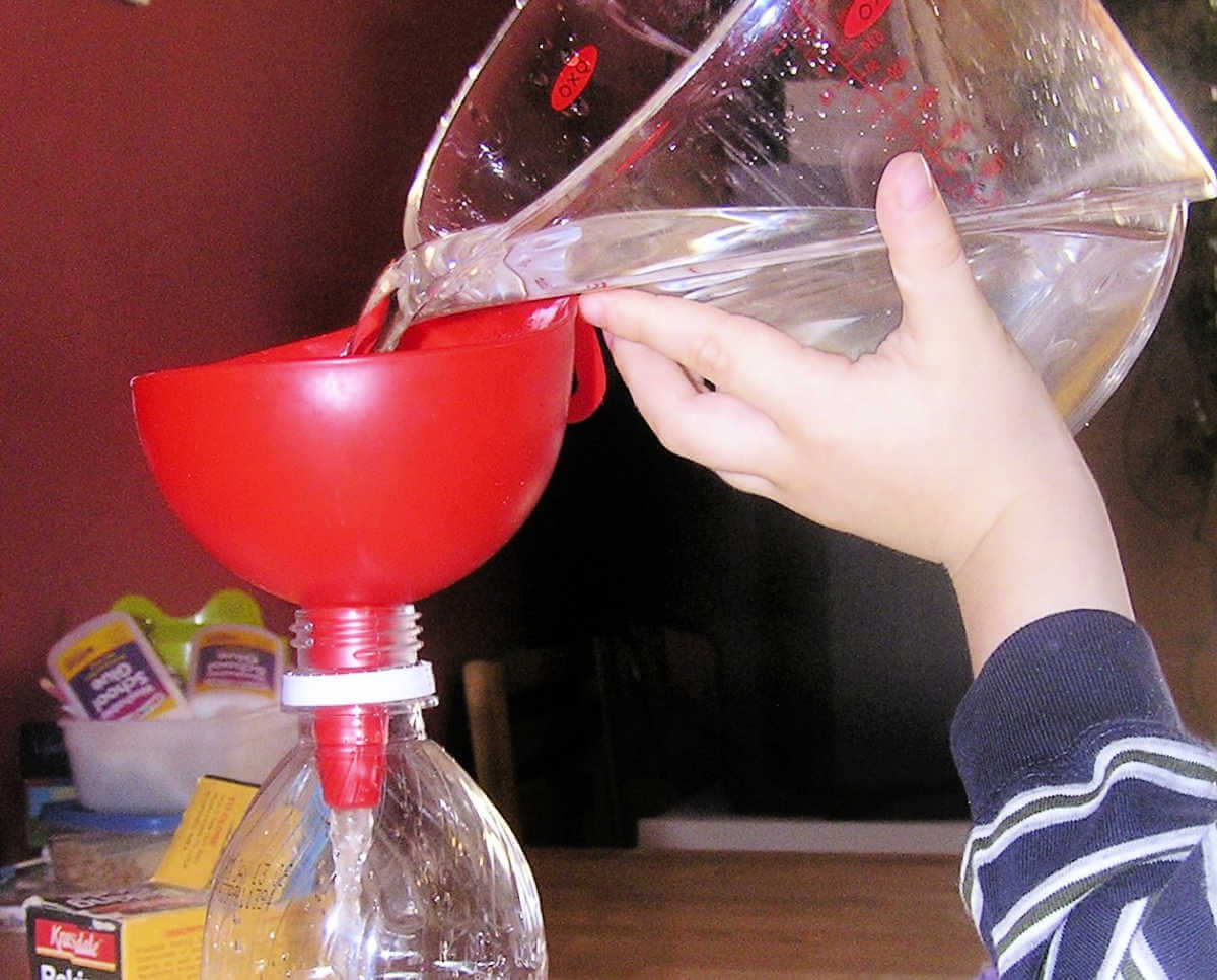 Child's hand pouring water from measuring cup into red funnel resting in bottle neck