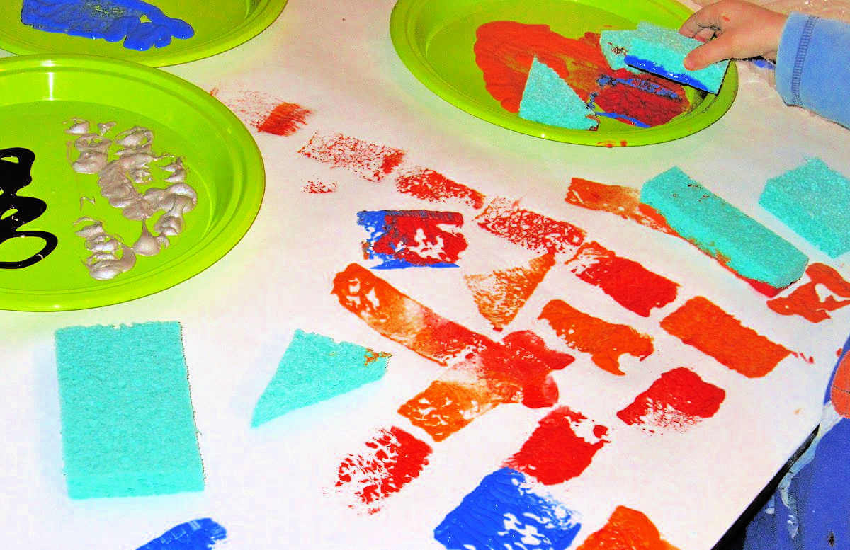 Child's hand placing sponge in paint surrounded by paper covered in paint, with sponges and paint on plates.