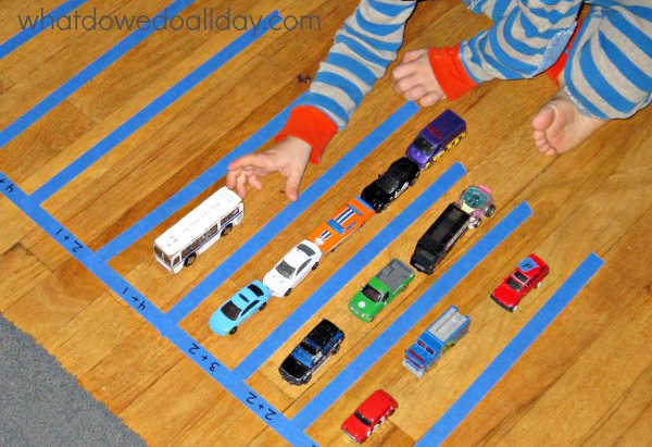 Child pushing toy vehicles into rows defined on the floor with blue tape.