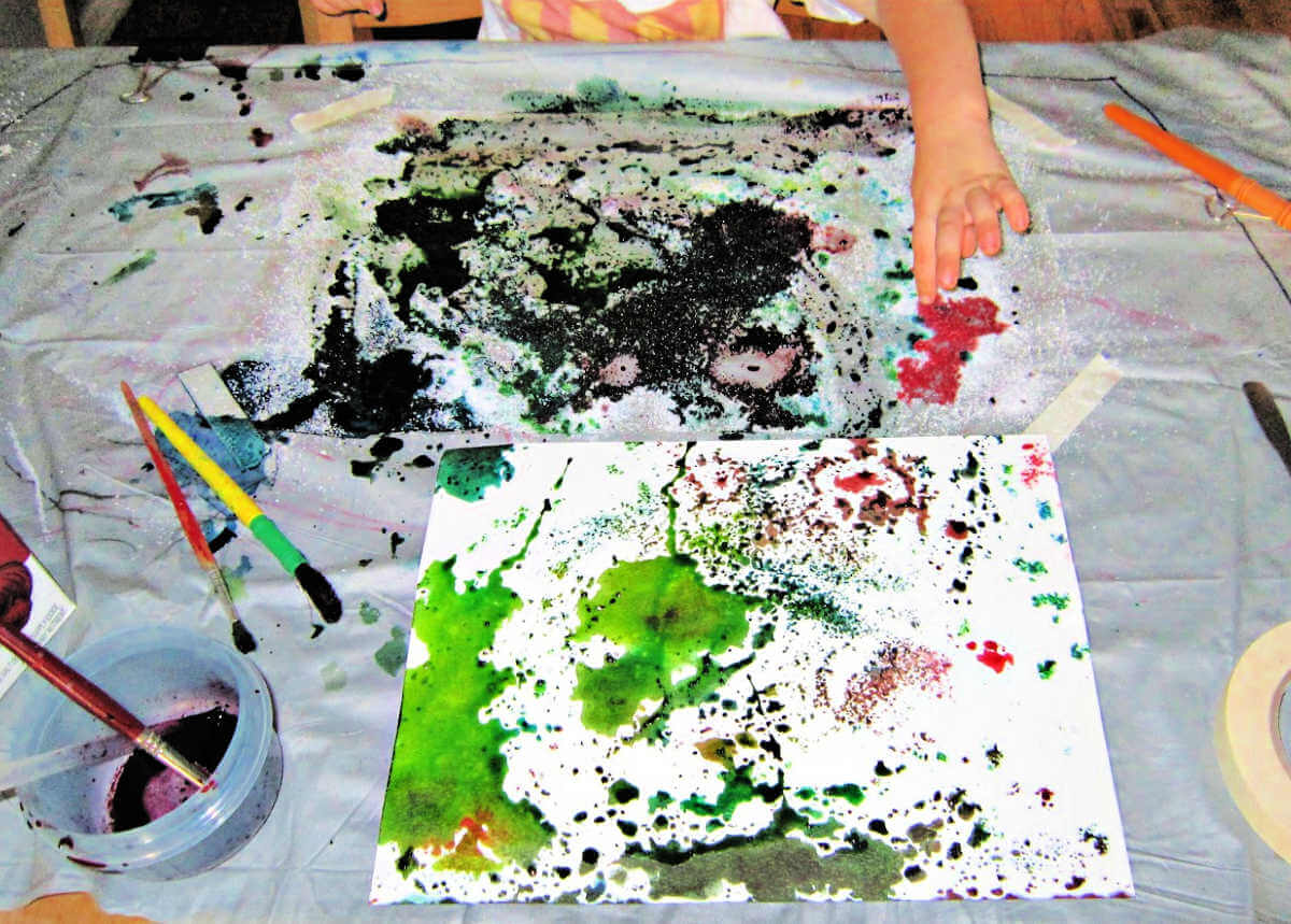 Child working on watercolor and salt art project at messy table covered in painting supplies