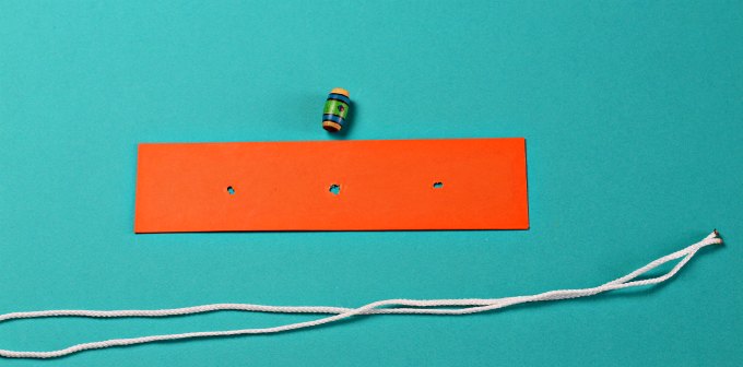 String and bead puzzle materials consisting of narrow orange board with three equidistant holes, white string, and one multicolored bead.